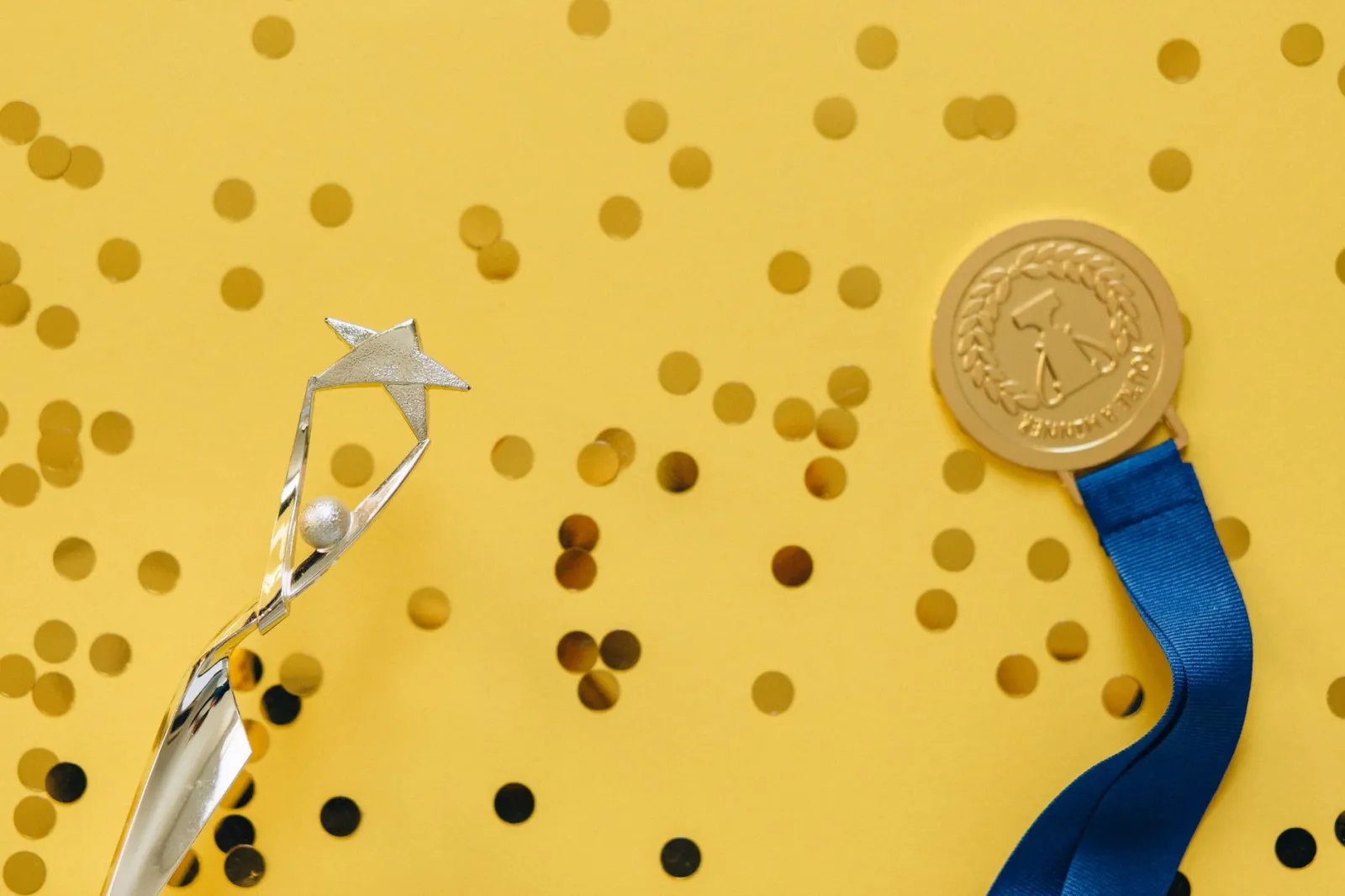 Golden medal and silver trophy with confetti on yellow surface, celebrating achievement.